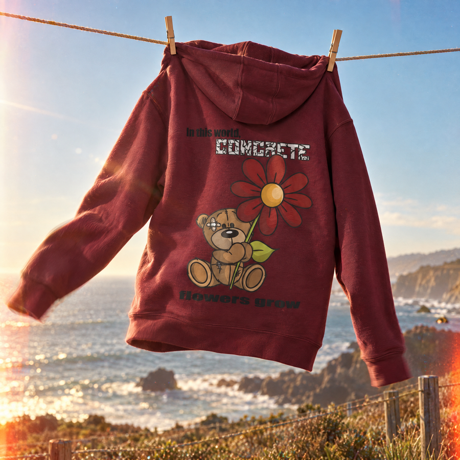 A maroon hoodie hanging from a clothesline outdoors, with a coastal sunset scene in the background. The hoodie features a graphic of a teddy bear holding a large red flower with the text 'in this world CONCRETE flowers grow'.
