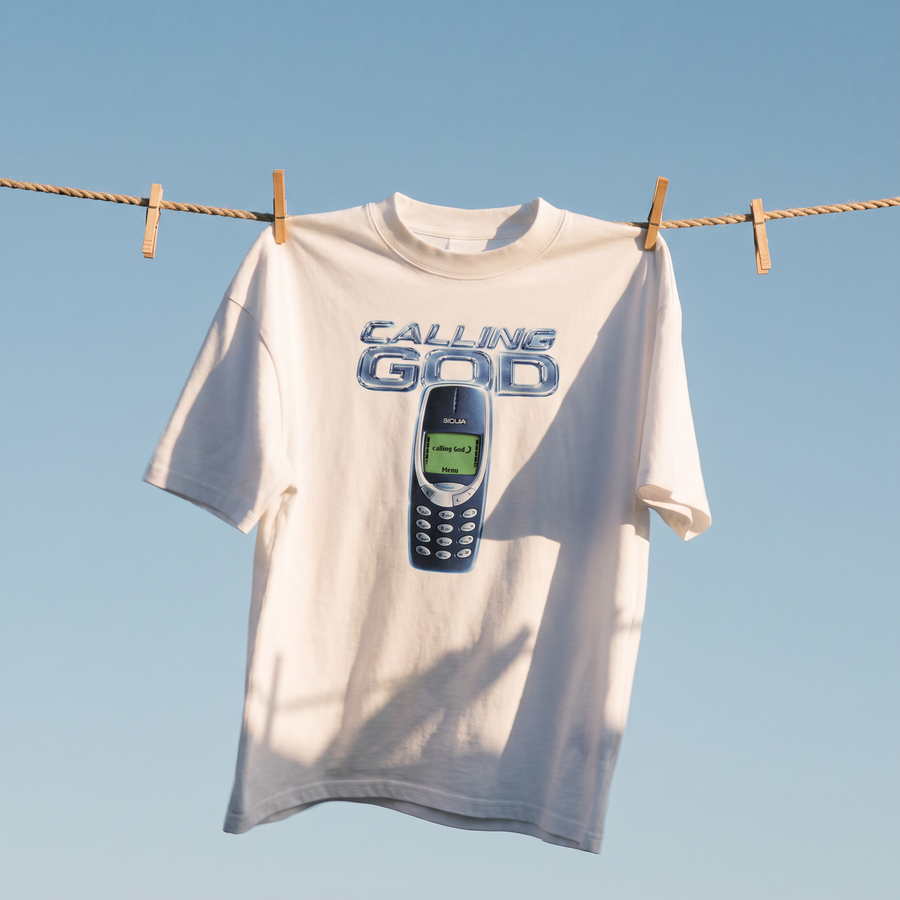 A white graphic t-shirt hanging on a rope clothesline with wooden clothespins against a clear blue sky. The shirt features a graphic print of a vintage blue mobile phone with the text 'CALLING GOD' in blue above it.