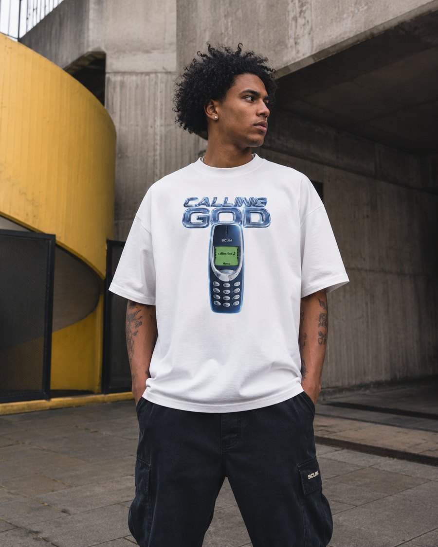 A man wearing a white graphic t-shirt with a blue and green 'Calling God' vintage mobile phone design, paired with black cargo pants, standing in an urban concrete environment.