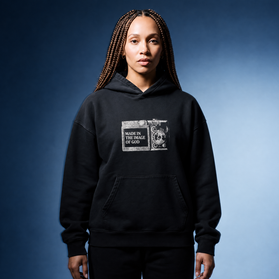 A woman with long braids stands against a blue gradient studio background wearing a black pullover hoodie with a distressed white graphic on the chest that reads 'MADE IN THE IMAGE OF GOD' alongside a skull illustration. She is also wearing black pants.