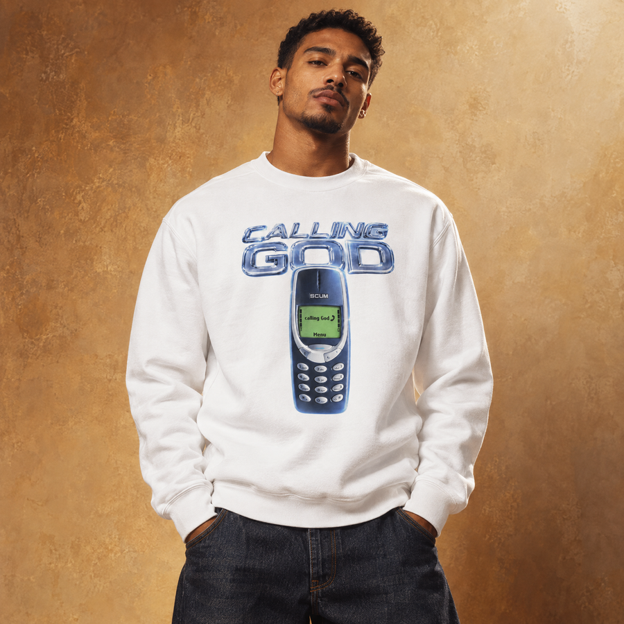 A man wearing a white crewneck sweatshirt with a blue graphic of a classic mobile phone and the text 'CALLING GOD' on the front, posed against a textured brown background.