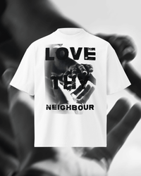 A white short-sleeve T-shirt with a large black graphic print on the front featuring the words 'LOVE THY NEIGHBOUR' over a hand image, shown against a blurred grayscale background of hands.
