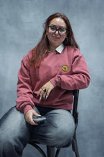 Person wearing the crimson sweater with a logo, sitting on a chair against a gray background
